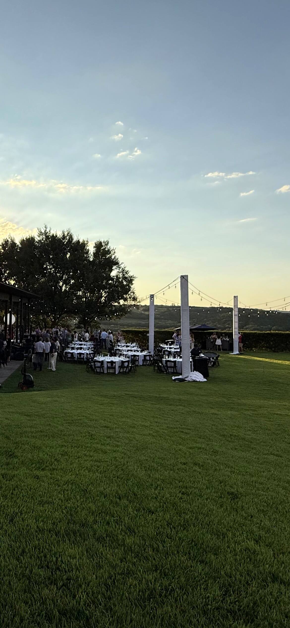A lawn set for dinner at sunset, white posts strung with lights